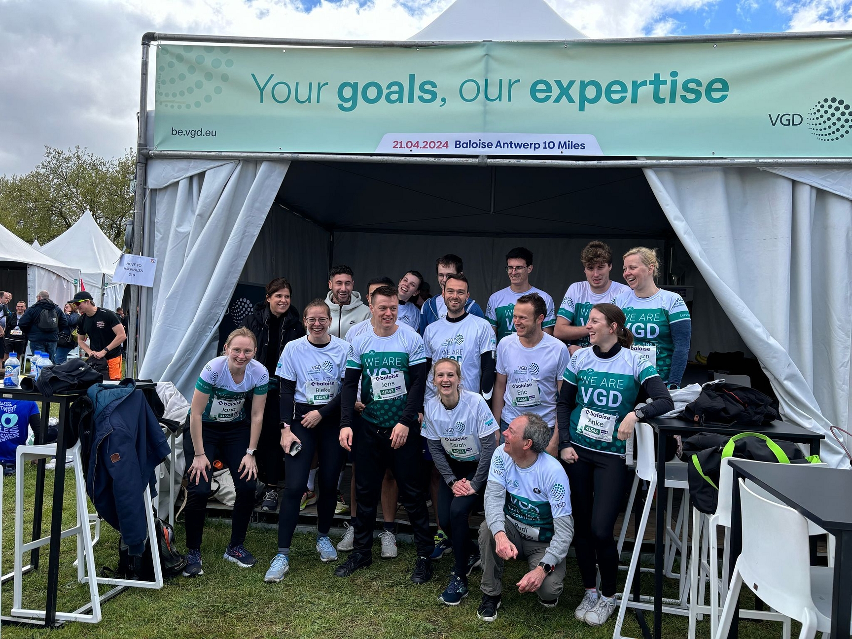 Je ziet een twintigtal collega's poseren voor een groepsfoto voor de VGD-tent in het Antwerp 10 Miles dorp. Enkelen staan rechtop, anderen zitten op hun knieën zodat iedereen voldoende plaats heeft. Ze dragen allen een loopshirt van VGD. Ze lachen naar elkaar en naar de camera.