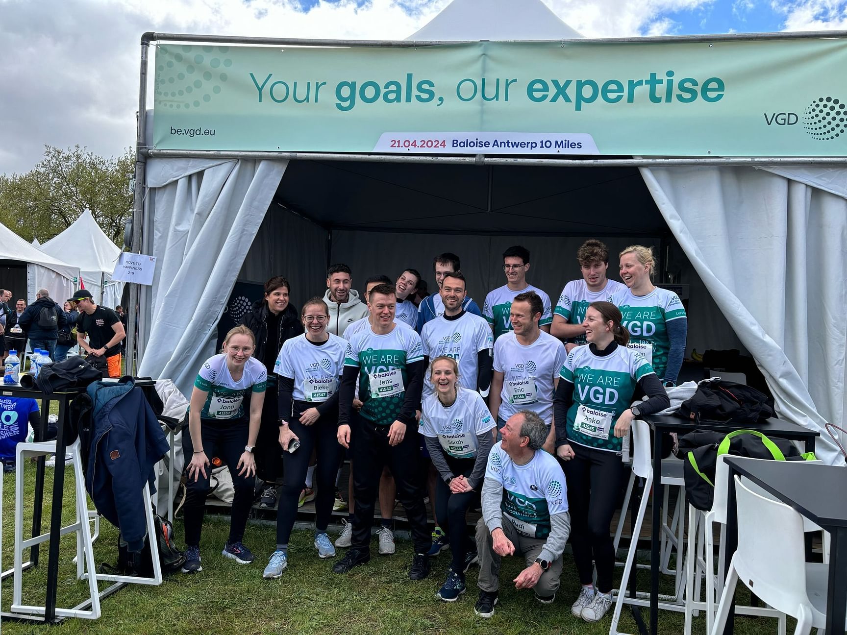 Je ziet een twintigtal collega's poseren voor een groepsfoto voor de VGD-tent in het Antwerp 10 Miles dorp. Enkelen staan rechtop, anderen zitten op hun knieën zodat iedereen voldoende plaats heeft. Ze dragen allen een loopshirt van VGD. Ze lachen naar elkaar en naar de camera.
