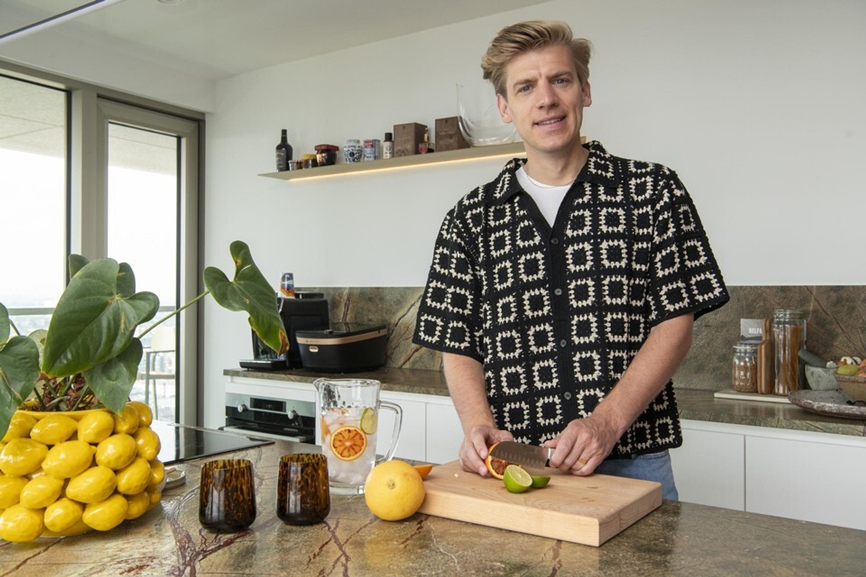 Je ziet de kok Michaël Sels tijdens een opname van een van zijn videorecepten. Hij staat in zijn keuken en kijkt in de camera. Het is duidelijk dat hij iets aan het uitleggen is over een gerecht dat hij aan het bereiden is. In zijn handen heeft hij een mes en een limoen vast en staat hij klaar om te beginnen snijden.
