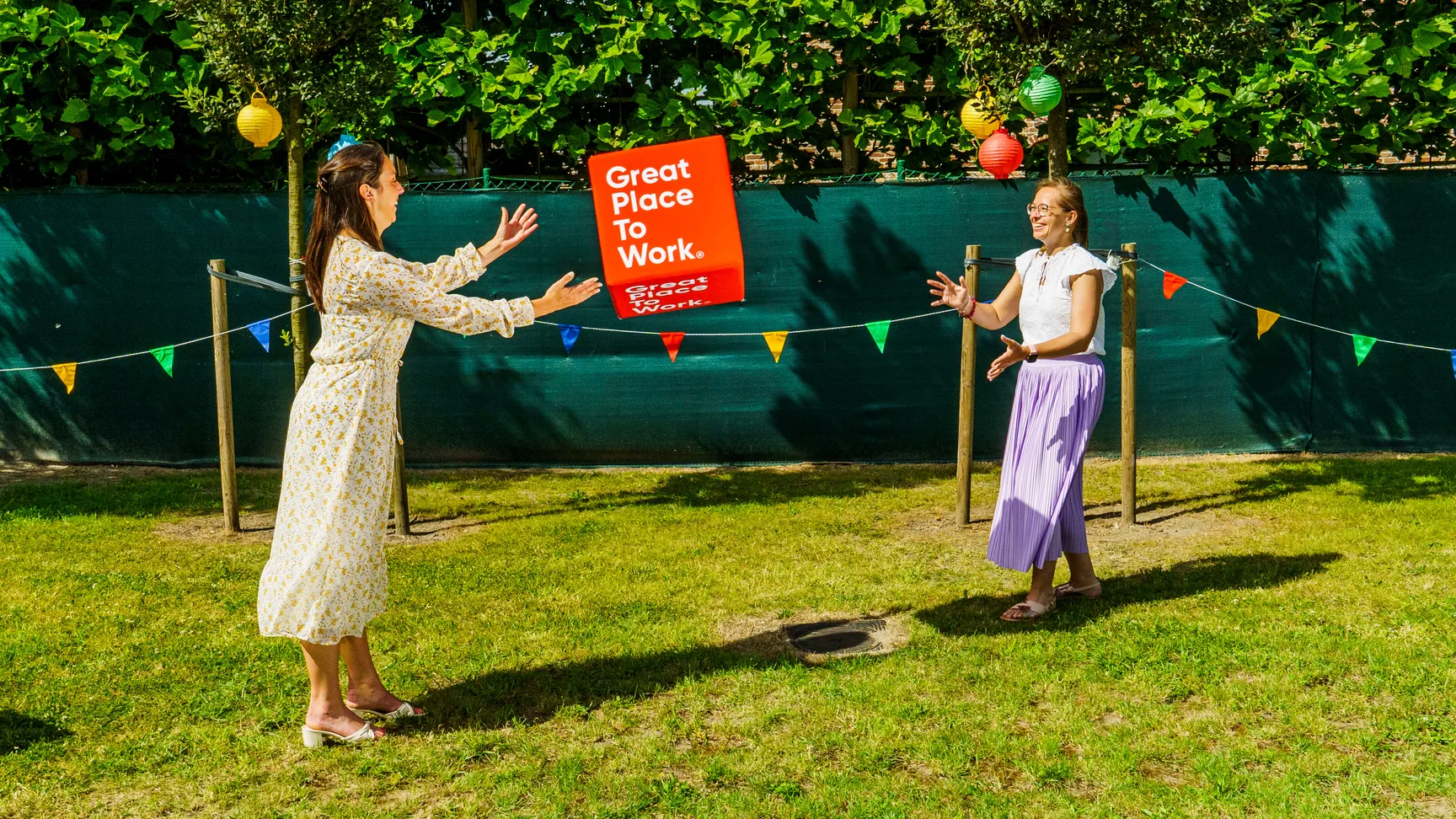 Op deze foto zie je onze collega's Bieke en Camille in een zomerse setting. Ze staan in een groene tuin waar kleurrijke vlaggetjes ophangen. Ze gooien een kubus naar elkaar. Op de kubus kan je het logo van Great Place to Work aflezen.