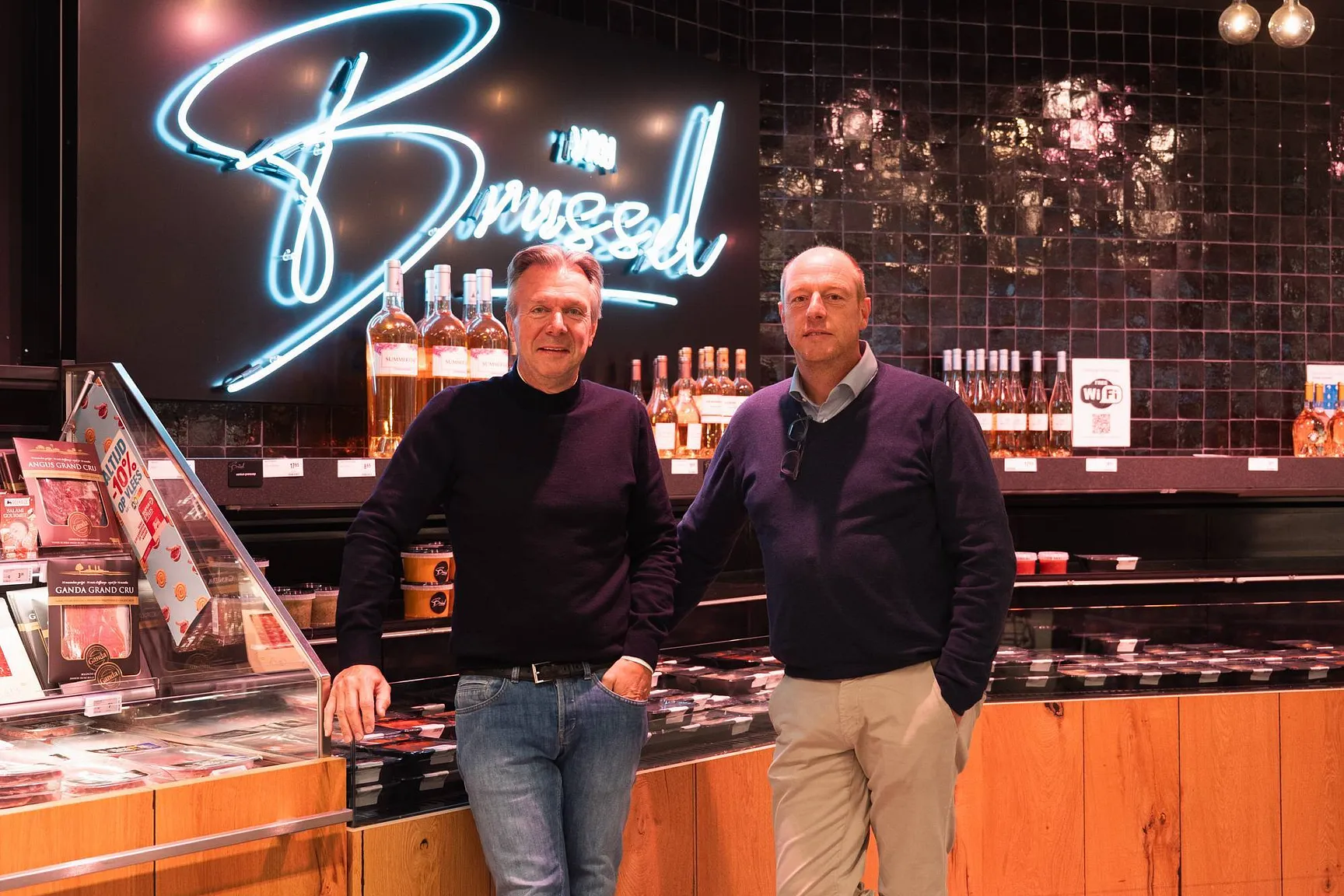 Twee personen poseren achter een toonbank in een winkel met verlichte decoratie op de achtergrond.