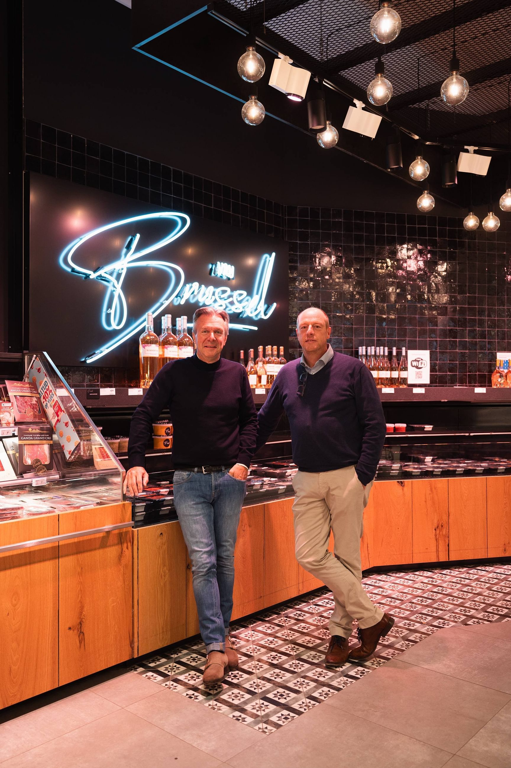 Twee personen poseren achter een toonbank in een winkel met verlichte decoratie op de achtergrond.