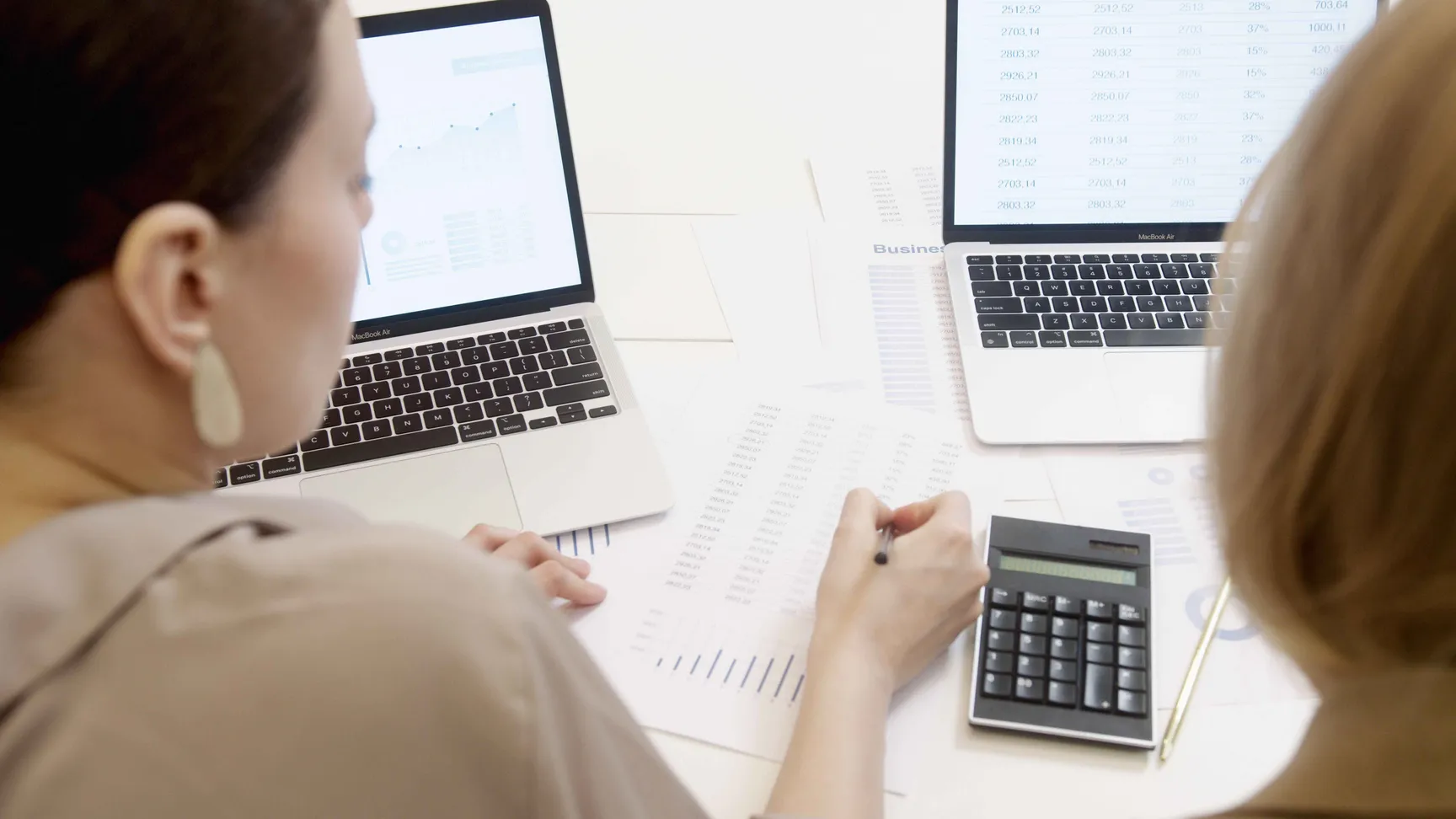 Een afbeelding van 2 personen die kijken naar financiële documenten. Op de bureau staan twee laptops en een rekenmachine.