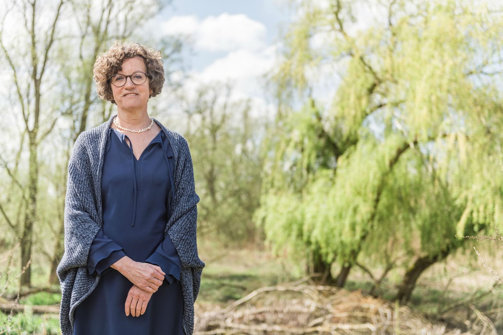 Een vrouw staat buiten in een groene omgeving met bomen en natuur op de achtergrond.