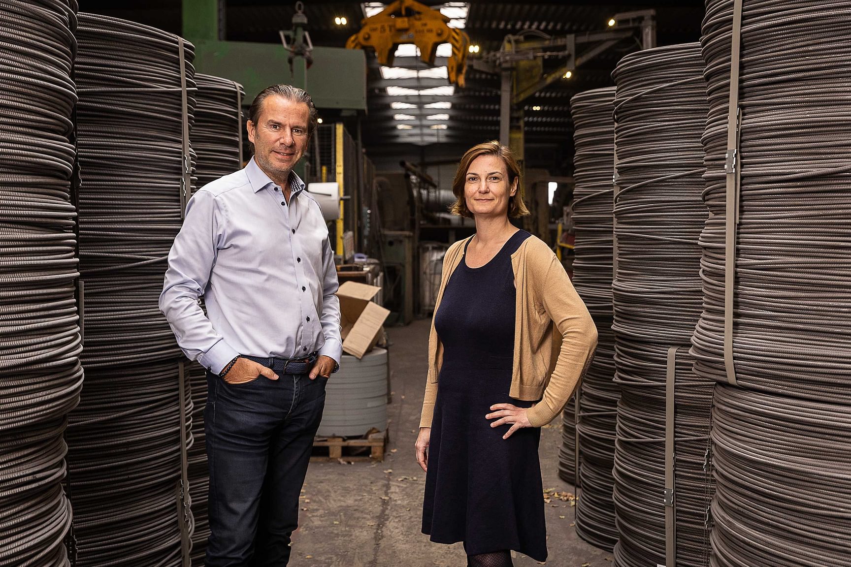 Twee personen staan in een productiehal tussen grote rollen materiaal op de achtergrond.