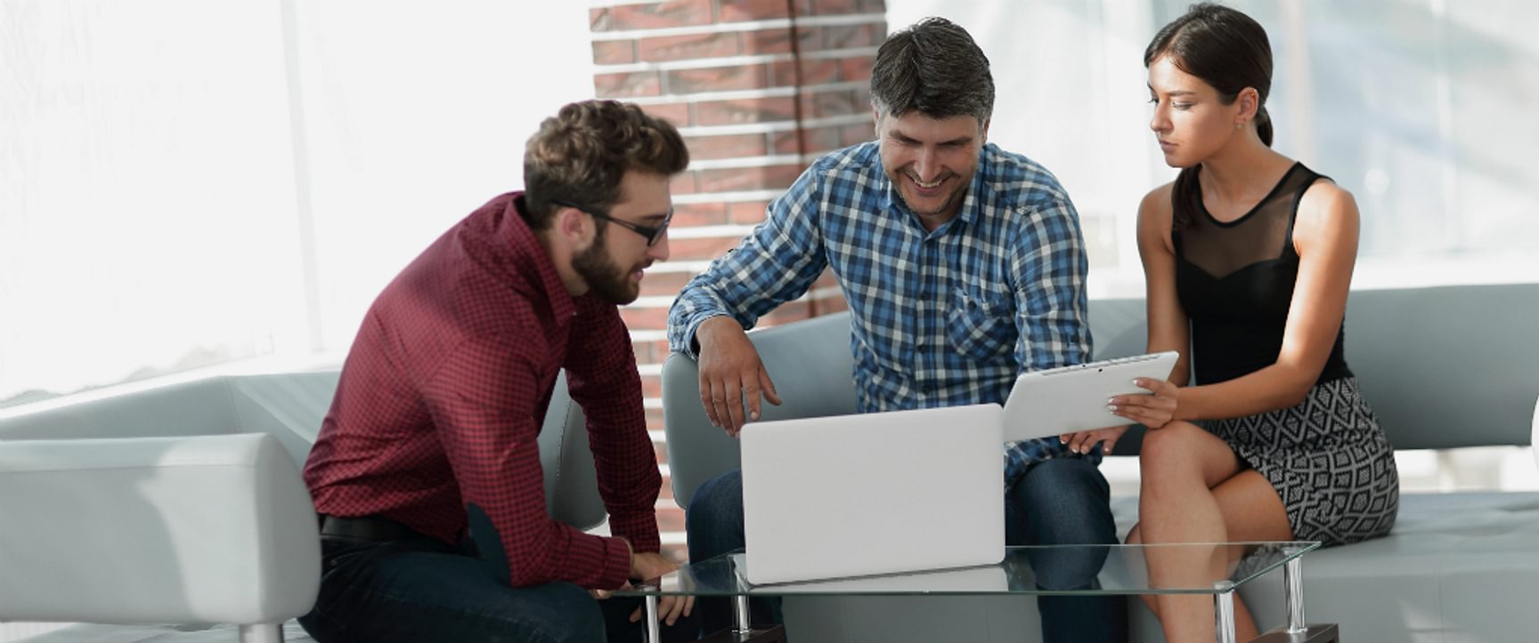 Drie personen bekijken samen een laptop en tablet tijdens een overleg op een zetel.