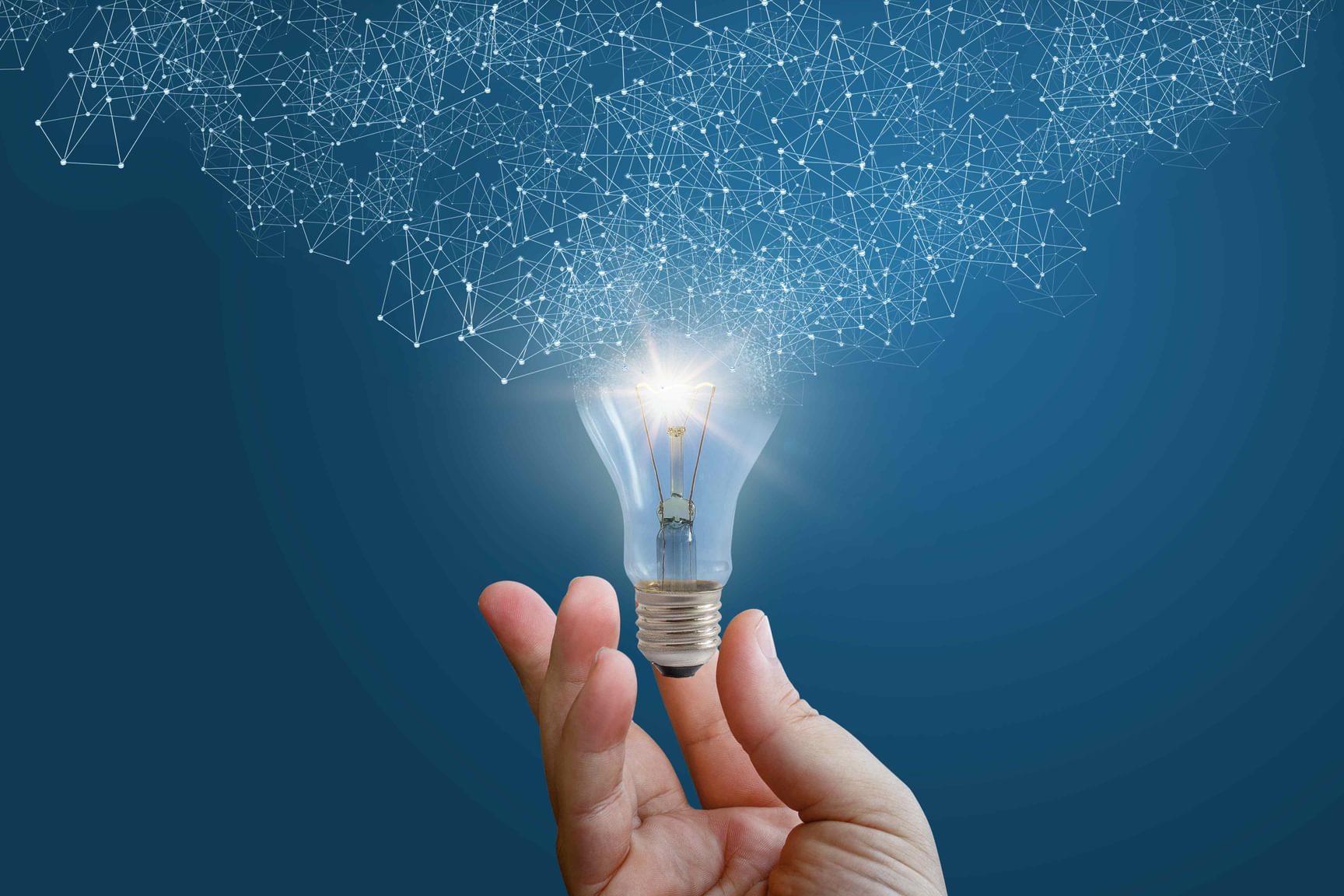 Hand houdt een brandende gloeilamp vast tegen een blauwe achtergrond, waaruit een netwerk van lichtpuntjes en lijnen omhoog straalt, als symbool voor ideeën, innovatie en technologie.