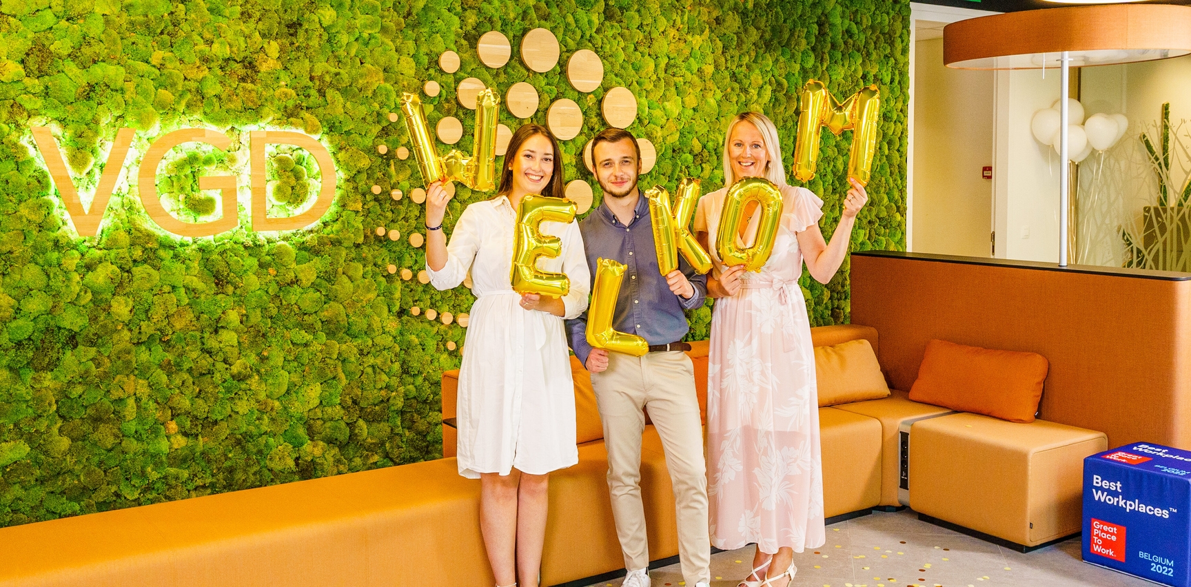 Collega's Sophie, Nils en Caroline houden gouden ballonnen vast in hun handen. De ballonnen vormen samen het woord welkom. Met een stralende glimlach poseren onze drie collega's voor onze prachtige groene moswand in onze werklplek in Wemmel. Hierop wordt het VGD logo heel mooi uitgelicht.