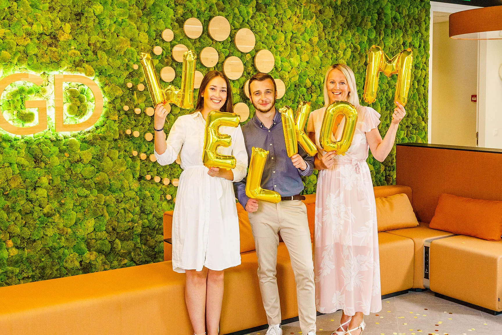 Collega's Sophie, Nils en Caroline houden gouden ballonnen vast in hun handen. De ballonnen vormen samen het woord welkom. Met een stralende glimlach poseren onze drie collega's voor onze prachtige groene moswand in onze werklplek in Wemmel. Hierop wordt het VGD logo heel mooi uitgelicht.