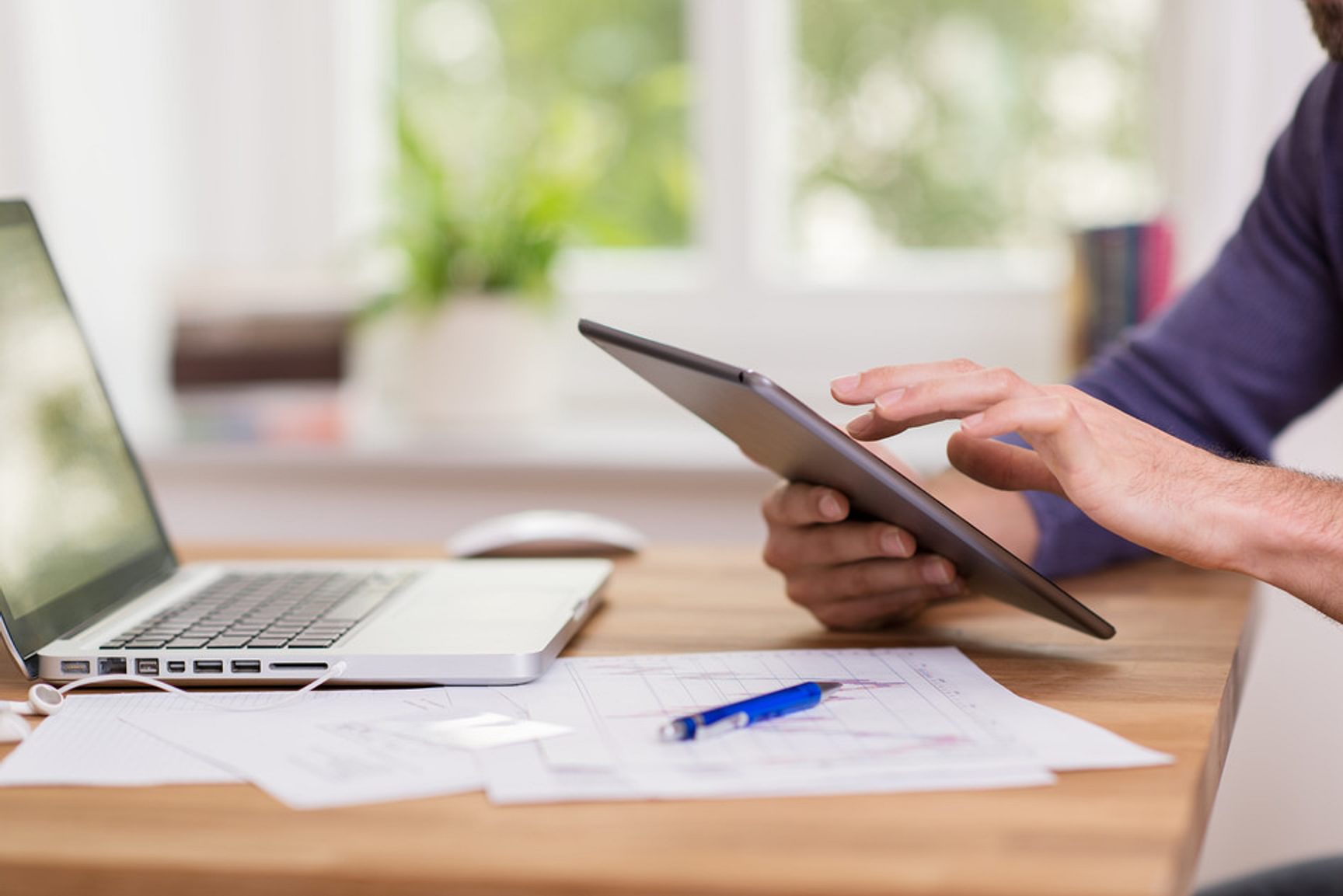 Persoon gebruikt een tablet aan een bureau met een laptop, pen en documenten.