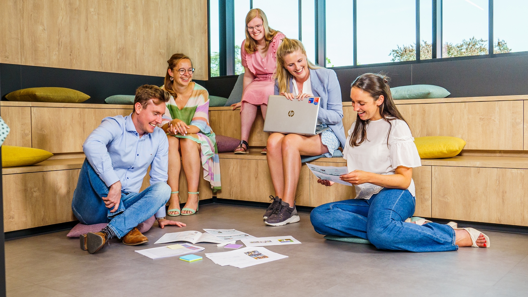 Een groep van vijf jonge professionals zit in een informele zithoek met houten banken en gekleurde kussens. Sommigen zitten op de grond, anderen op de banken, terwijl ze brainstormen. Ze dragen kleurrijke kledij en luisteren naar elkaar. De sfeer is ontspannen en creatief, met grote ramen die natuurlijk licht binnenlaten.