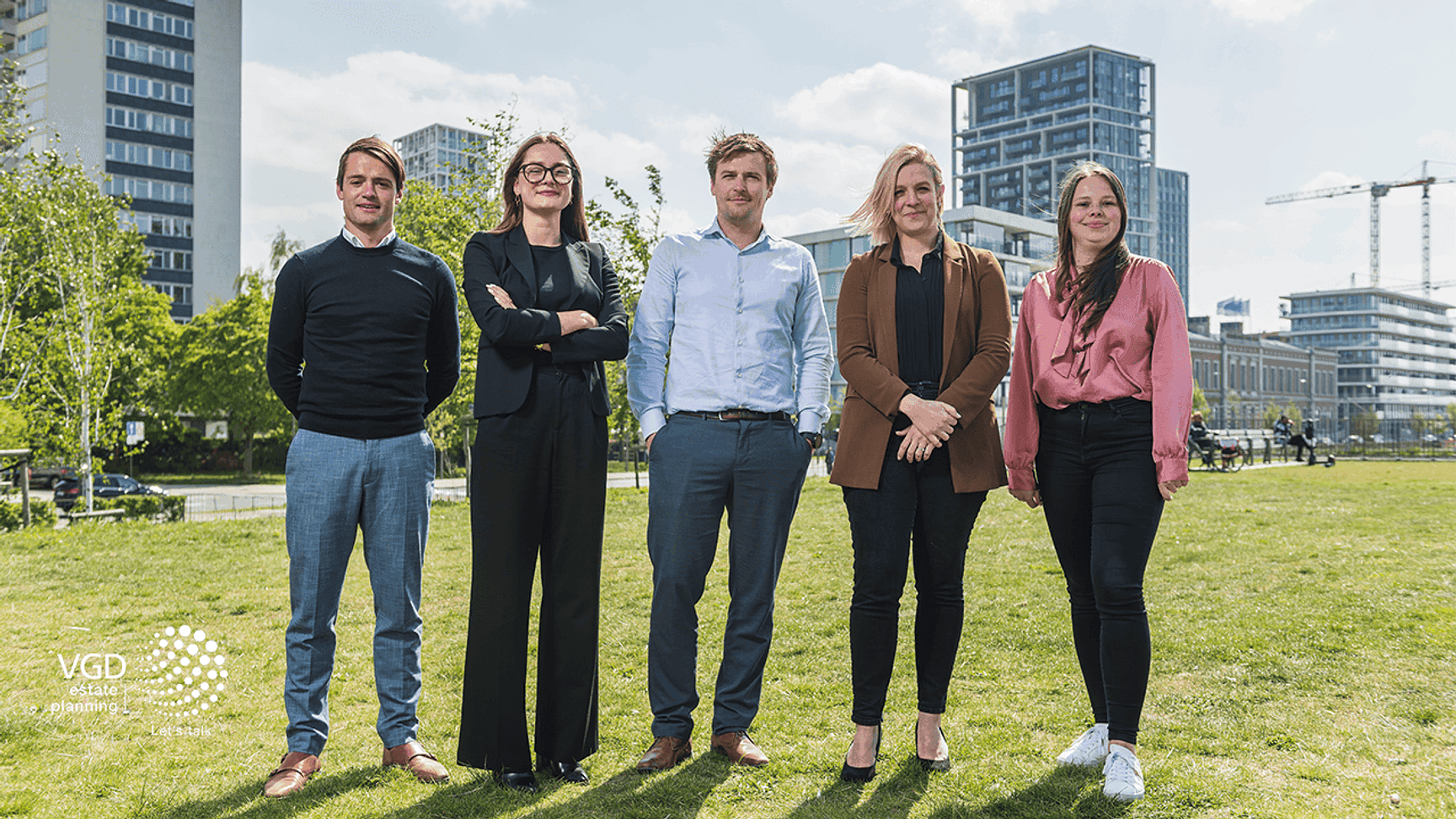 Groep van vijf personen staat samen buiten op een grasveld met moderne gebouwen op de achtergrond.