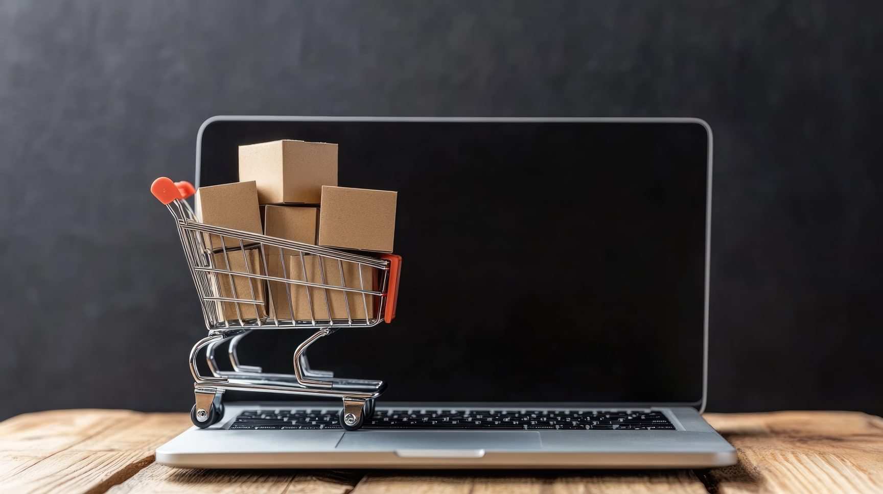 Een laptop dat op een tafel staat met daarop een miniatuur winkelwagen gevuld met kartonnen dozen.