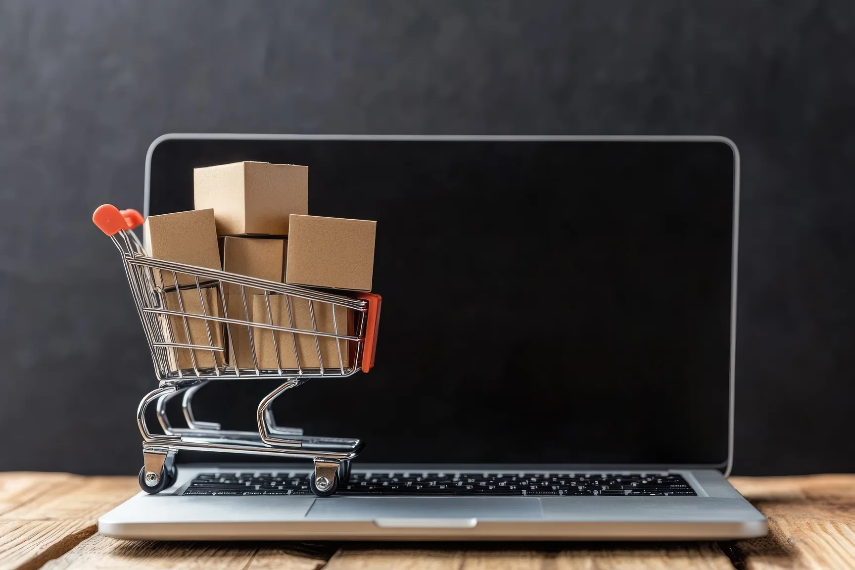 Een laptop dat op een tafel staat met daarop een miniatuur winkelwagen gevuld met kartonnen dozen.