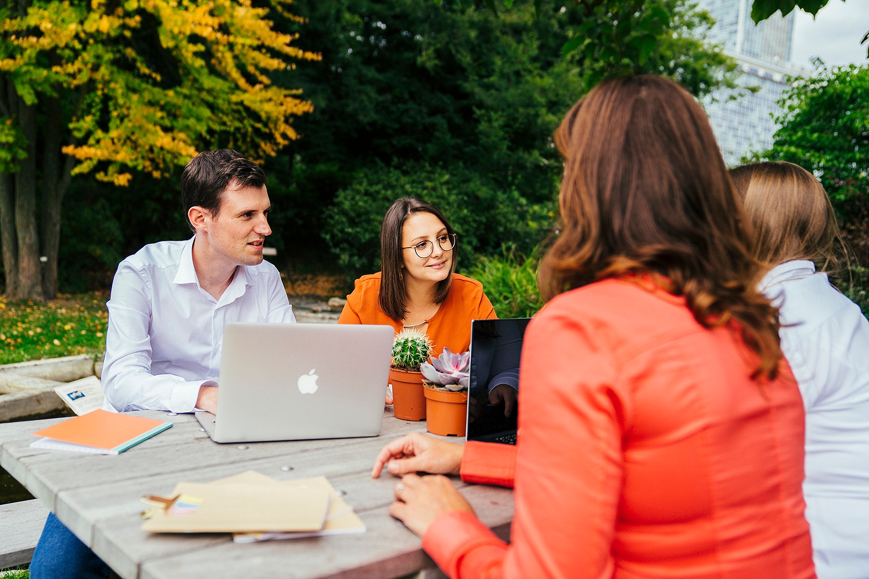 Je ziet vier collega's zitten aan een picknickbank buiten. Op de tafel staat een laptop open, liggen enkele notitieboekjes open. De collega's overleggen met elkaar.