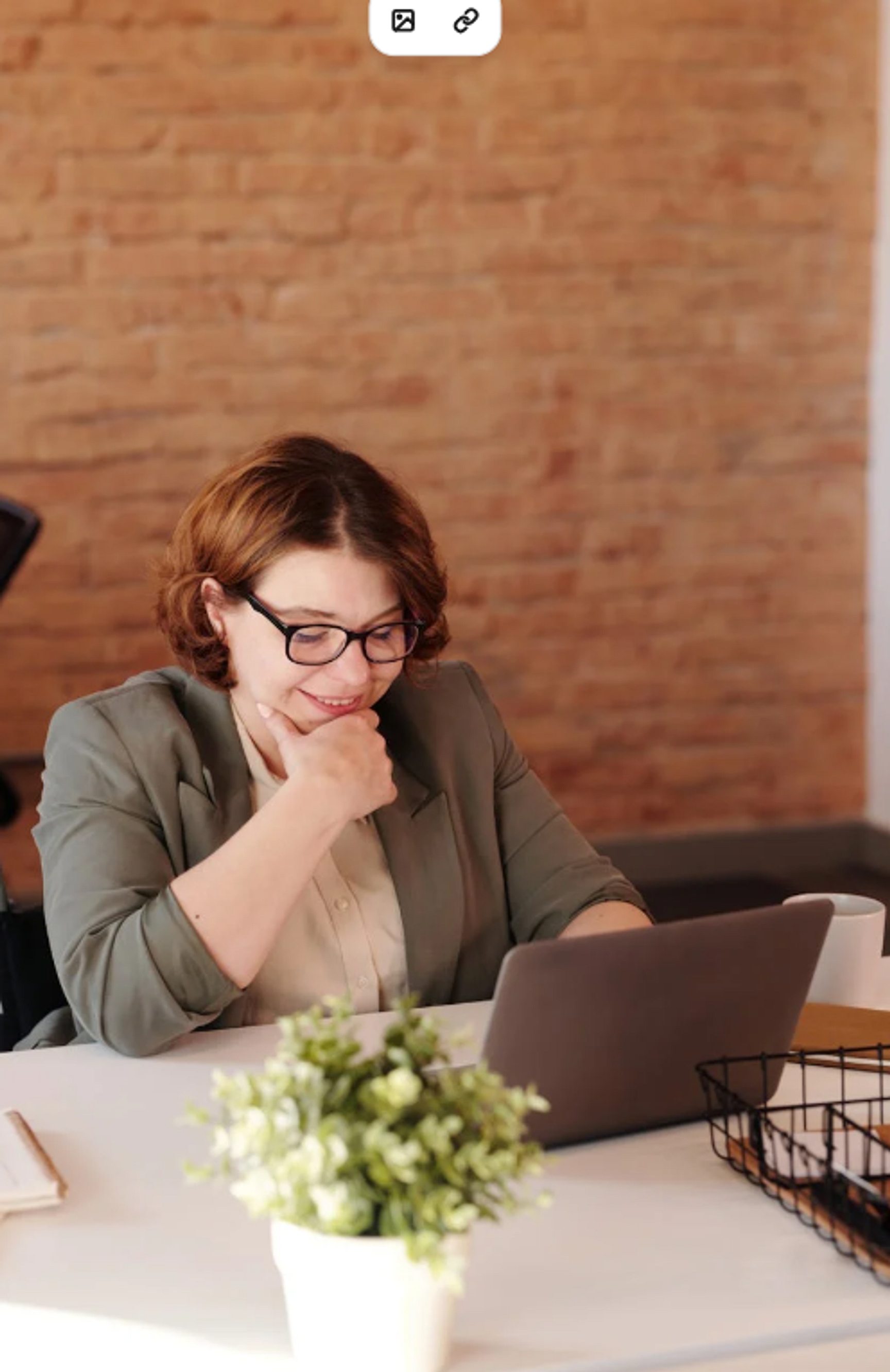 Je ziet een dame lachend naar haar computerscherm kijken. De vrouw heeft kort bruin haar en draagt een zwarte bril. In de voorgrond staat er een klein plantje.