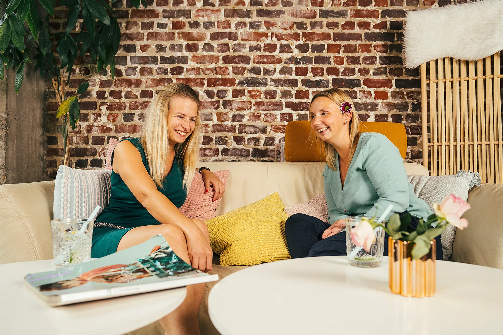 Op de foto zitten onze collega's Magdalena en Jennifer samen op een comfortabele sofa. Ze babbelen en lachen met wat er verteld wordt.  De pastelkleurige kussens geven de foto een warme en gezellige uitstraling.