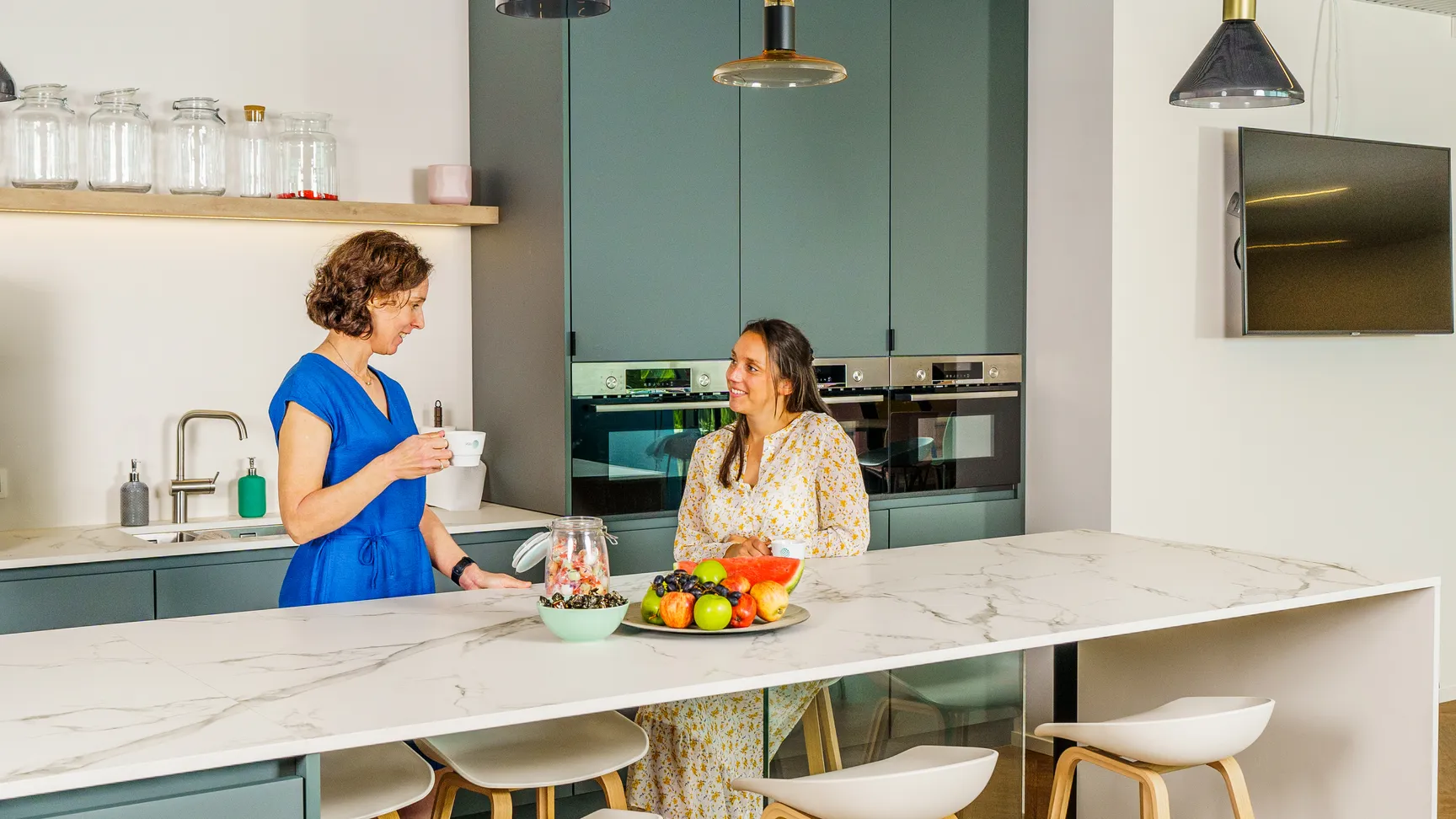 Je ziet onze collega's Camille en Yolande aan het keukeneiland van onze moderne keuken in werkplek Melle. Yolande staat recht en houdt een kopje koffie vast. Camille zit neer en praat met Yolande. Voor hen staat een fruitschaal gevuld met kleurrijk fruit.