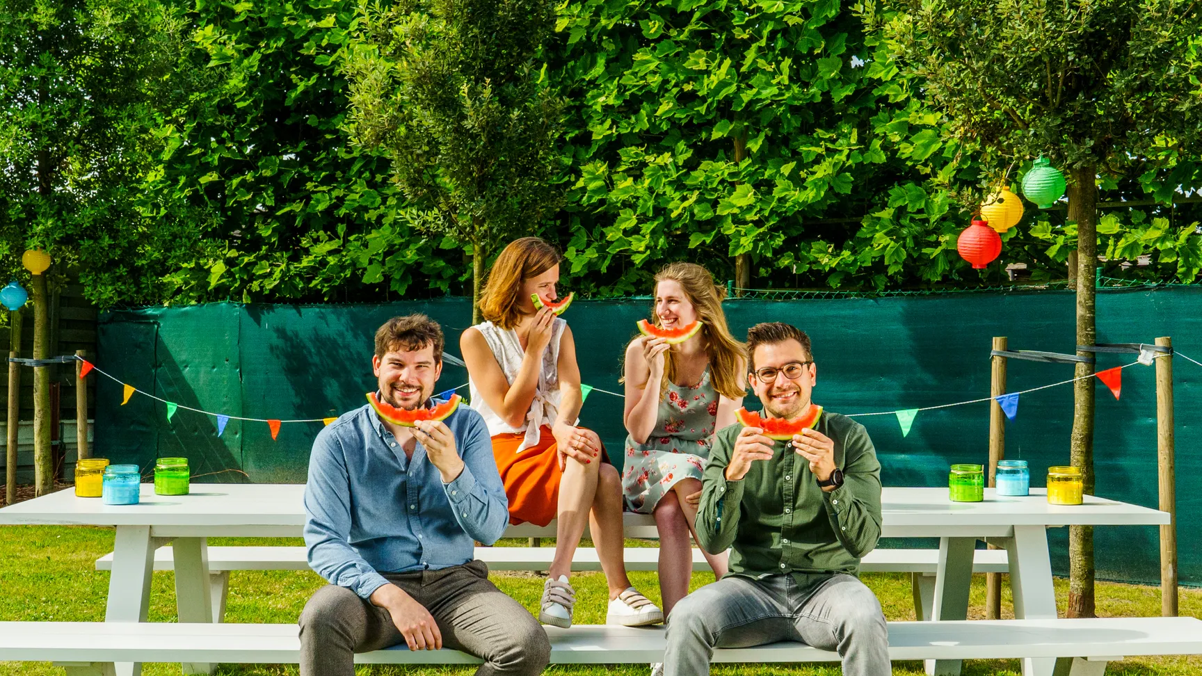 Je ziet vier collega's zitten op een picknick bank in de tuin van onze werkplek Melle. De twee mannen kijken lachend in de camera en houden een snede watermeloen vast in hun handen. De twee dames kijken naar elkaar en houden de watermeloen als een smiley voor hun gezicht.