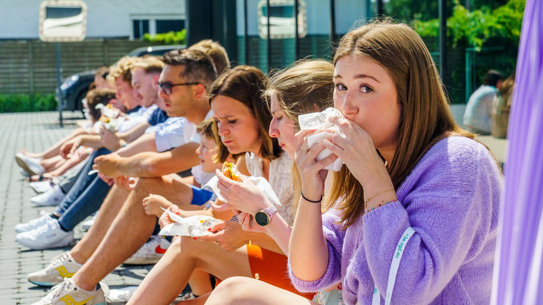 Deze foto werd genomen tijdens een traktatie van een van onze partners. Op deze zonnige dag zie onze collega's genieten van een burger. Ze zitten op een rijtje naast elkaar uit het vuistje te eten. De collega dichtst bij de camera merkt net op dat de fotograaf een foto neemt wanneer ze een hap van haar burger neemt. De foto geeft een gezellige en informele sfeer weer.