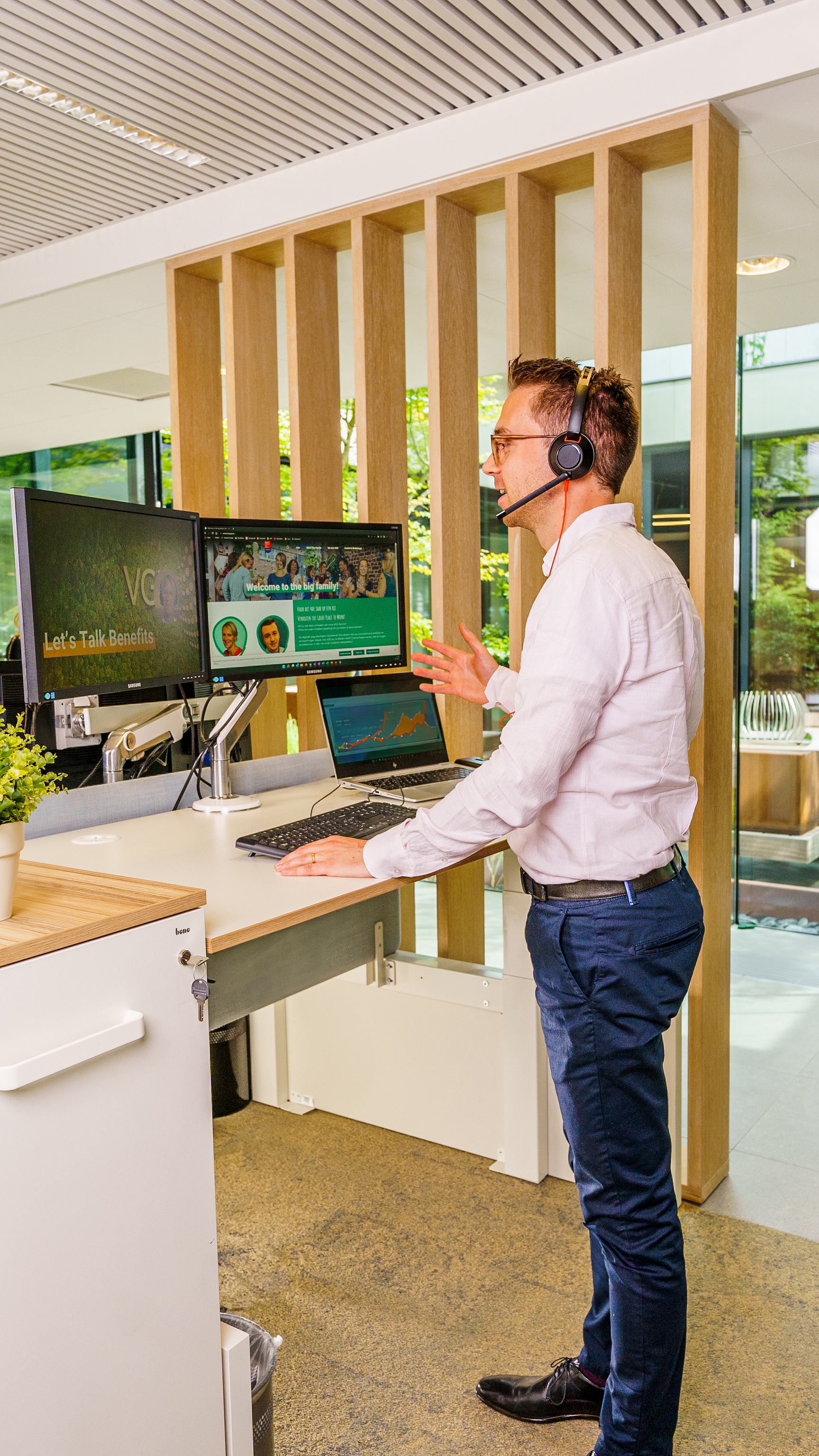 Onze collega Koen staat aan een staande bureau te werken. Hij belt met zijn koptelefoon en legt iets uit. Hij gebruikt zijn handen om iets te vertellen aan een collega in een Teams video call.