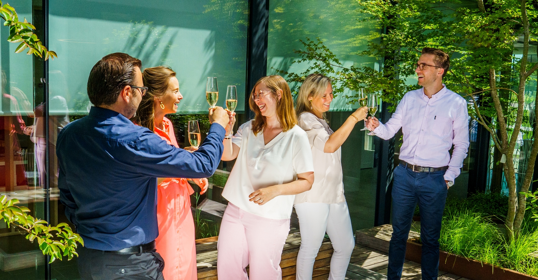 Je ziet 5 collega's in de binnentuin van onze werkplek in Wemmel. Ze houden allen een glas bubbels in de lucht klaar om met elkaar te klinken. Het zonnetje en de glimlachen zorgen ervoor dat er een zeer fijne en toegankelijke sfeer heerst.