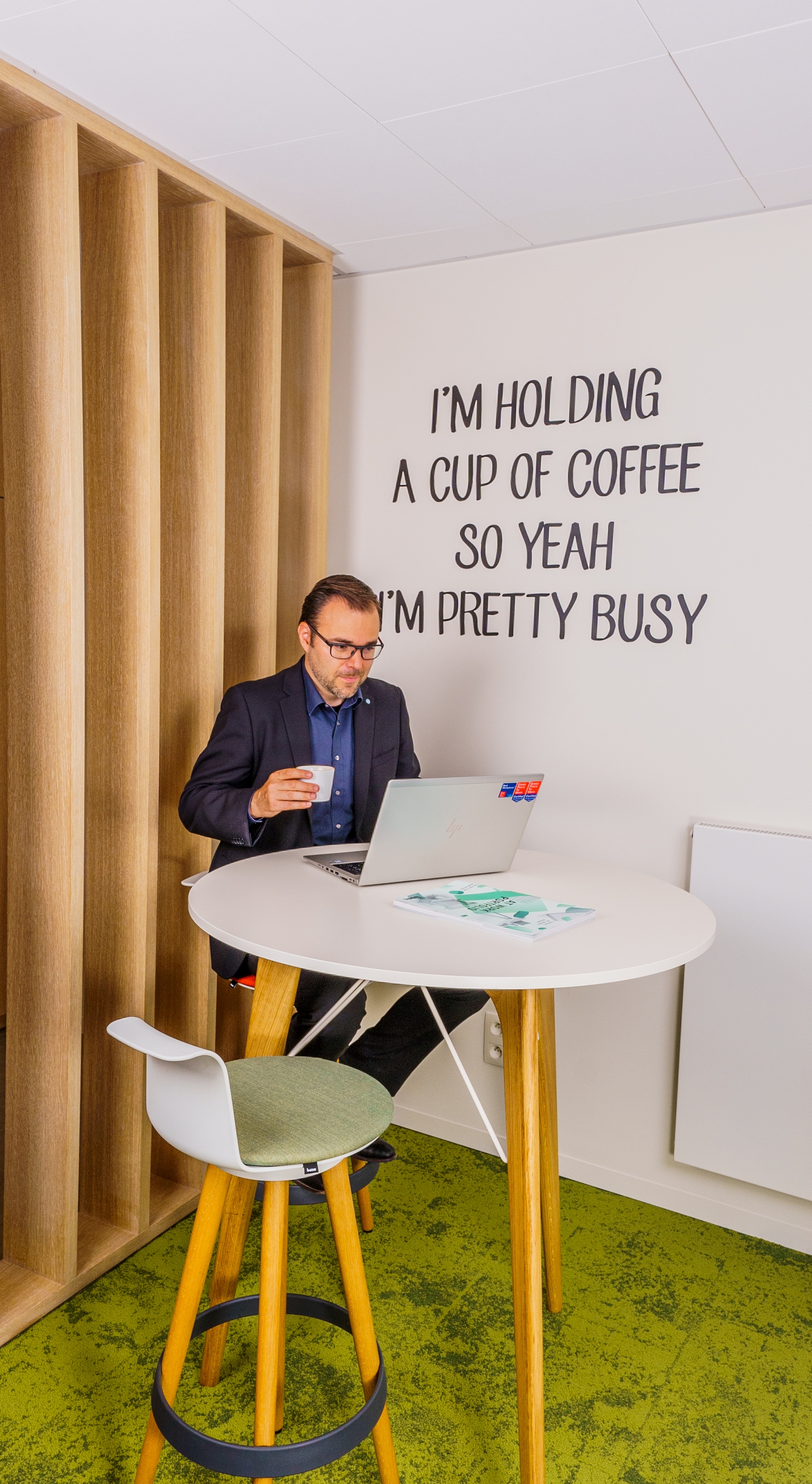 Je ziet collega Erik aan een hoge tafel zitten in onze werkplek in Wemmel. Hij houdt een kopje koffie vast en leest iets op zijn laptop. Op de muur achter hem zie je de quote hangen "I'm holding a cup of coffee. So yeah i'm pretty busy"