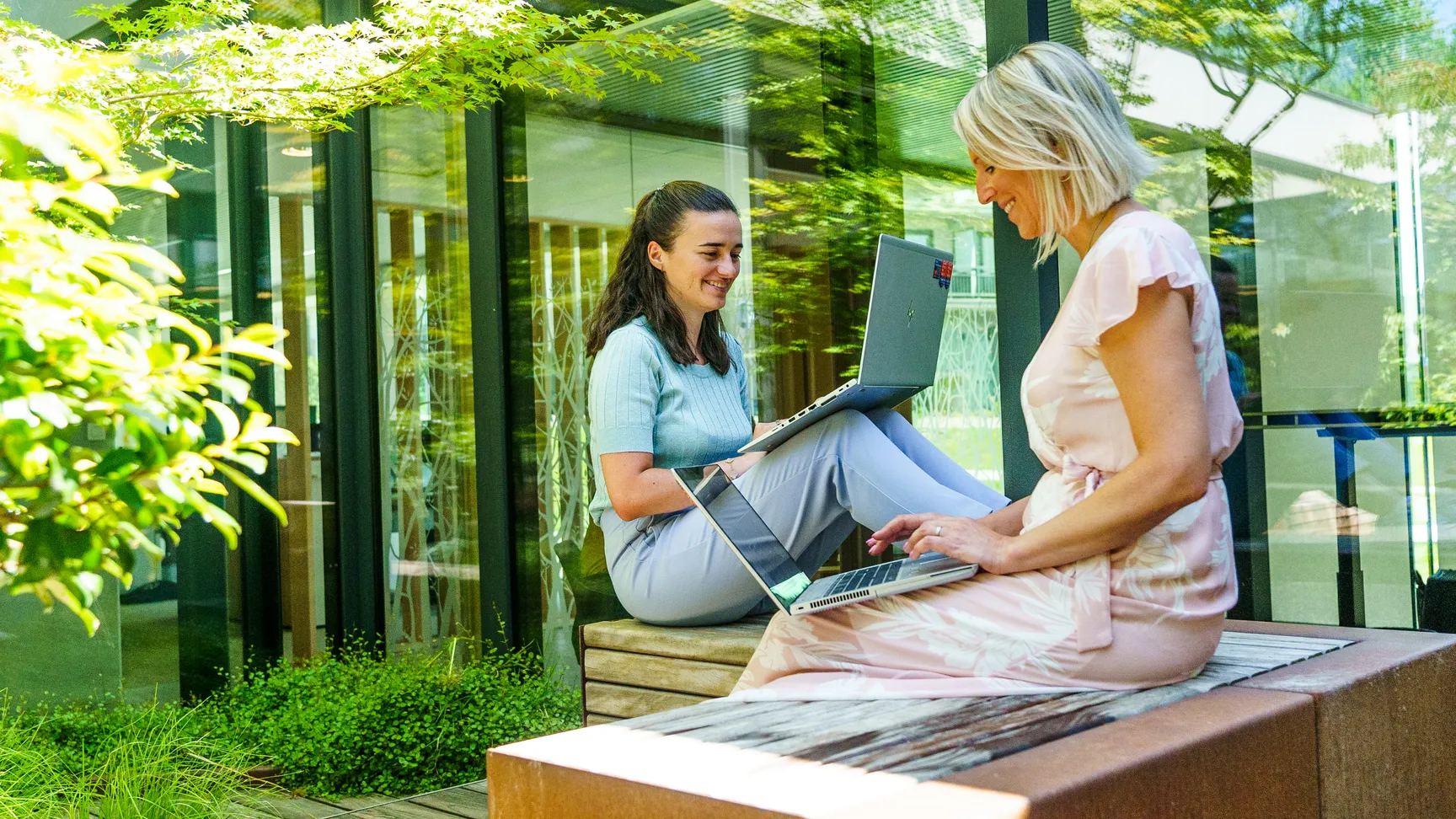 Twee collega's zitten naast elkaar op een bankje in de buitentuin van onze werkplek in Wemmel. Ze zijn beide vrolijk aan het werk op hun laptop.