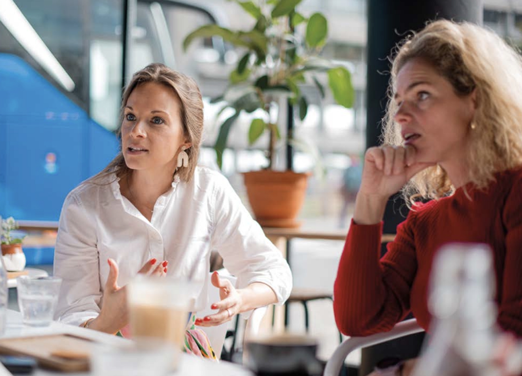 Twee vrouwen zitten aan een tafel in een lichte, moderne ruimte. De vrouw links draagt een witte blouse en is in gesprek met een persoon buiten beeld. De vrouw rechts, gekleed in een rode trui, luistert aandachtig met haar hand tegen haar kin naar de persoon buiten beeld. Op de tafel staan een waterglas en een latte macchiato, en op de achtergrond is een grote plant te zien.