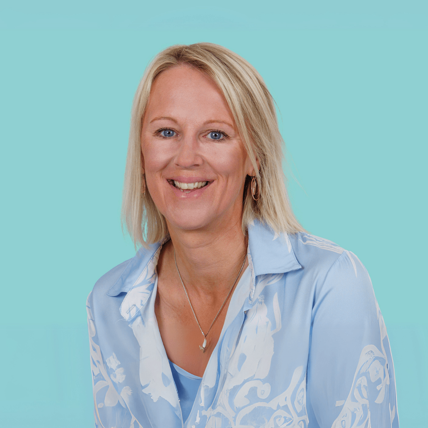 Caroline kijkt met een warme glimlach in de camera. Ze heeft blond haar en draagt een licht blauwe blouse. De foto straalt warmte en energie uit door de complimentariteit met de zachte blauwe achtergrond.